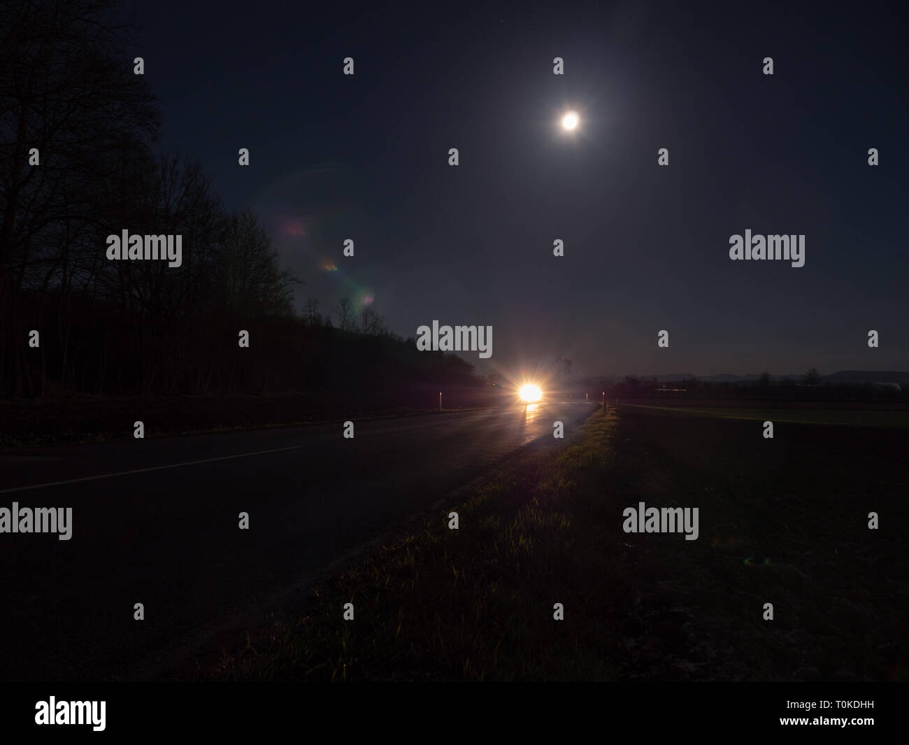 Route de campagne sinueuse éclairé par les phares d'une voiture avec la pleine lune et paysage mystérieux Banque D'Images