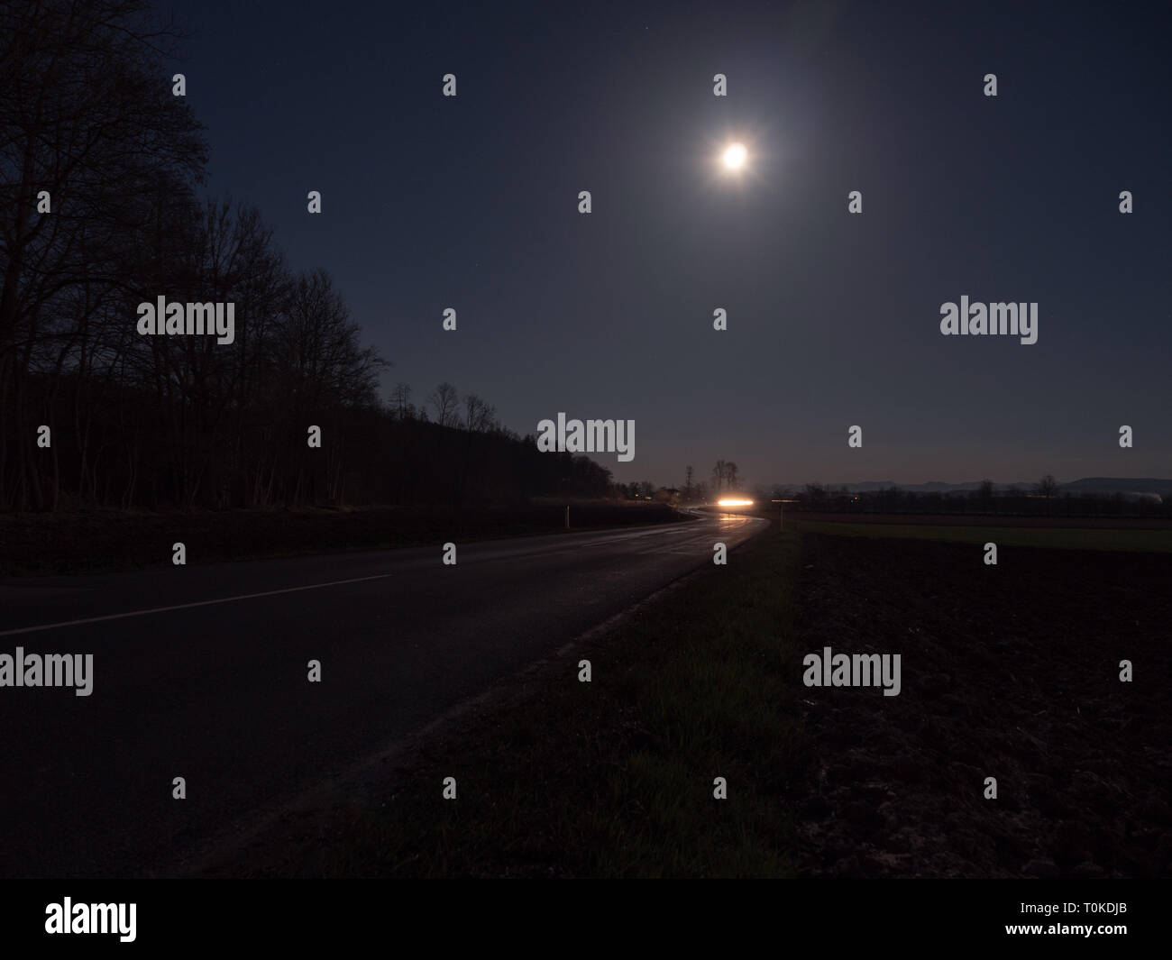 Route de campagne sinueuse éclairé par les phares d'une voiture avec la pleine lune et paysage mystérieux Banque D'Images