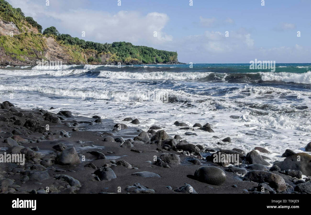 Plage de sable noir : un rouleau, le barattage surfez polit des pierres de lave et des dépôts de sable noir sur une plage dans les îles hawaïennes. Banque D'Images