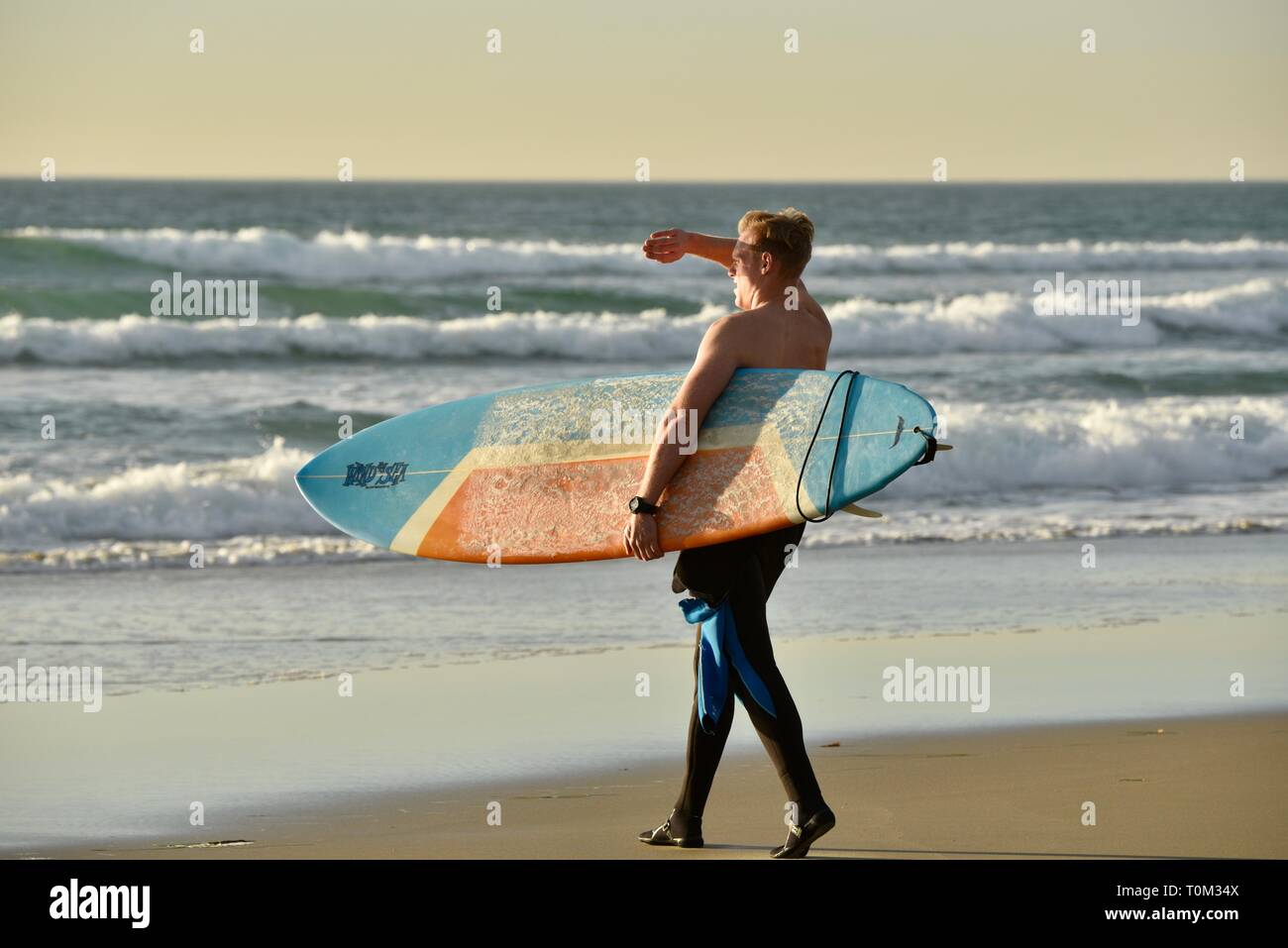 Homme séduisant à marcher en direction de l'eau de l'océan au coucher du soleil avec une planche de surf sous le bras et le blocage du bras avec Sun, Pacific Beach, San Diego, CA, USA Banque D'Images