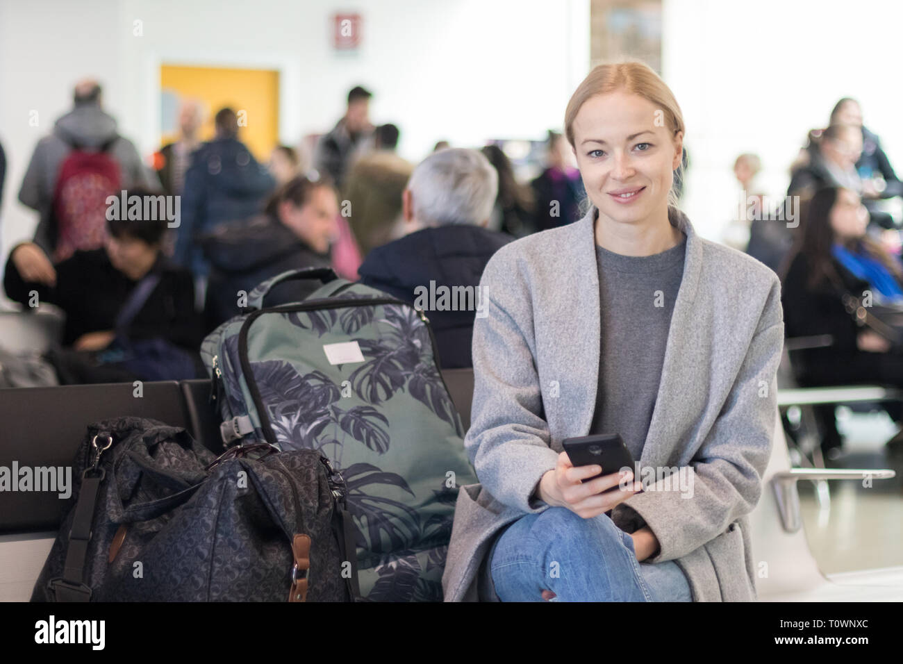 Cheerful female traveler smiling at camera, lors de la lecture sur son téléphone portable en attendant de monter à bord d'un avion à l'embarquement à l'aéroport Banque D'Images
