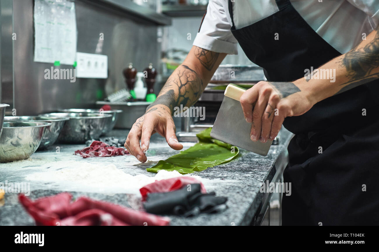 Recette secrète. Photo recadrée des mains du chef de tatouages pâte de coupe sur la table de la cuisine avec de la farine pour pâtes italiennes faites maison. Banque D'Images