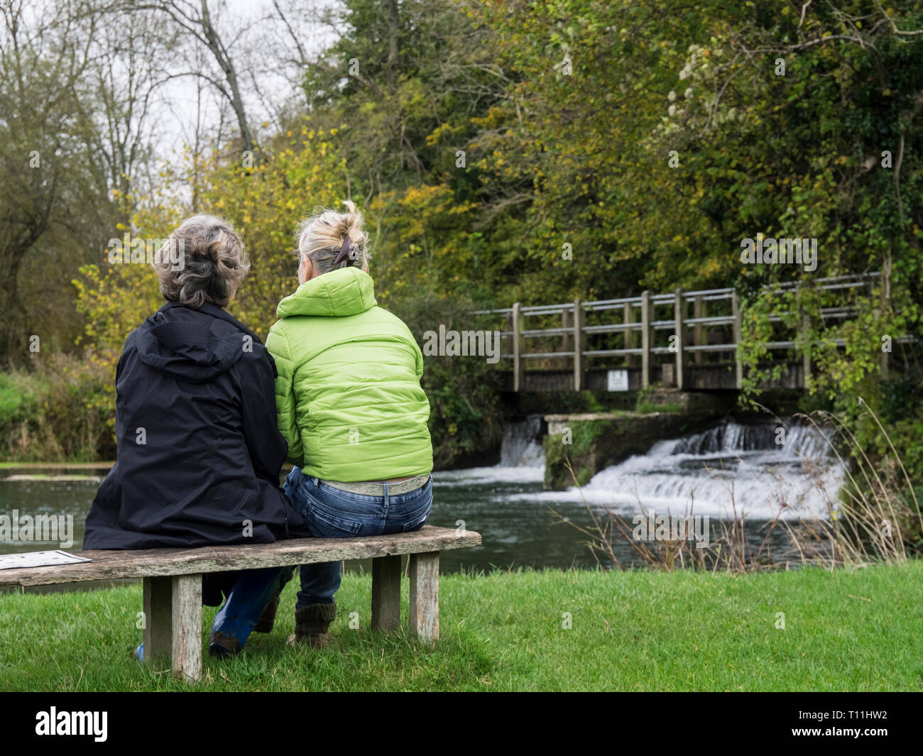 Vue arrière d'une mère et sa fille chat portant des vestes, assis sur un banc au bord d'une petite cascade et Weir en arrière-plan Banque D'Images