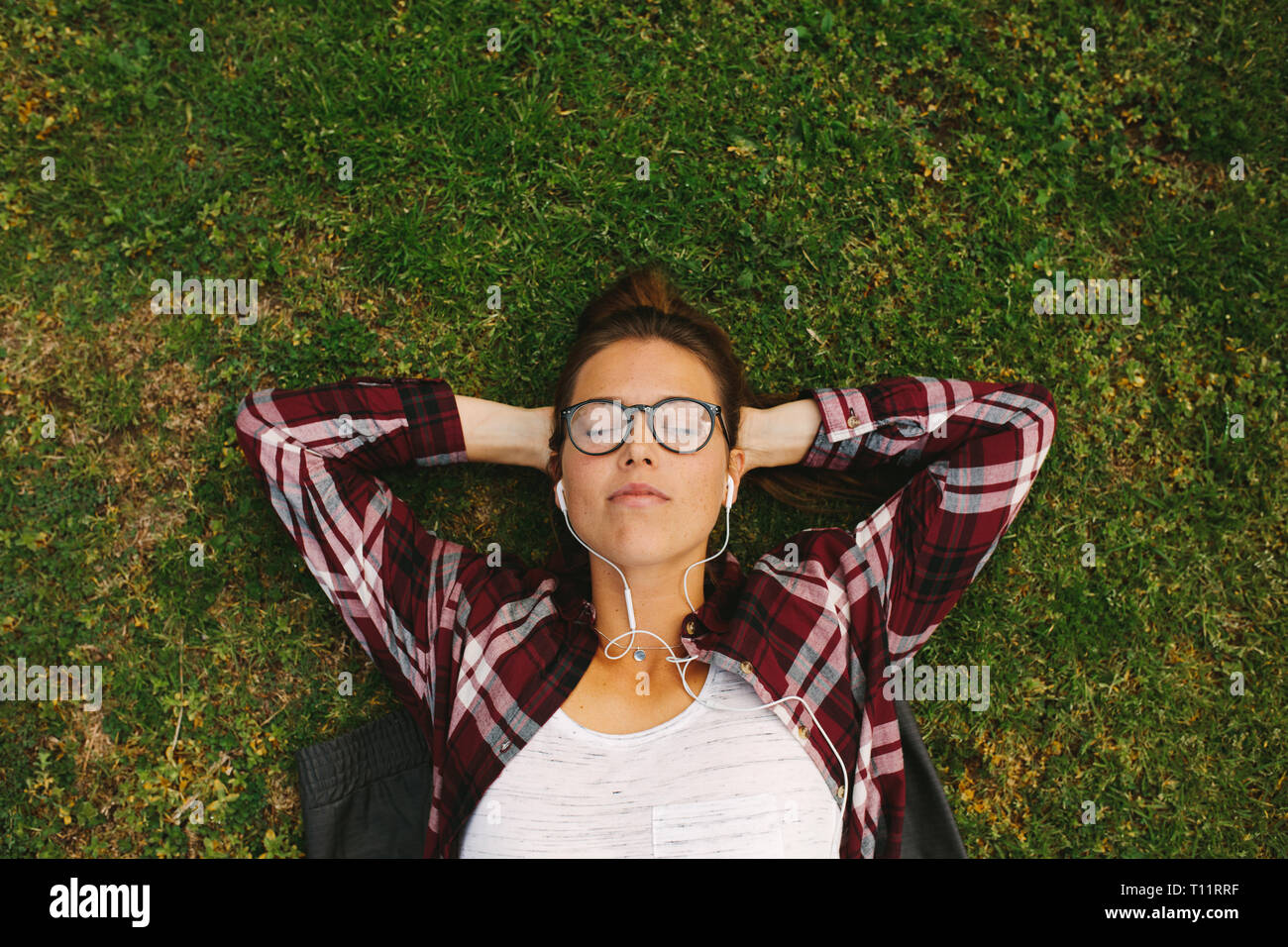 Top View of young woman wearing eyeglasses écoutez de la musique avec des écouteurs. Female student lying on grass avec les mains derrière la tête et de détente à col Banque D'Images