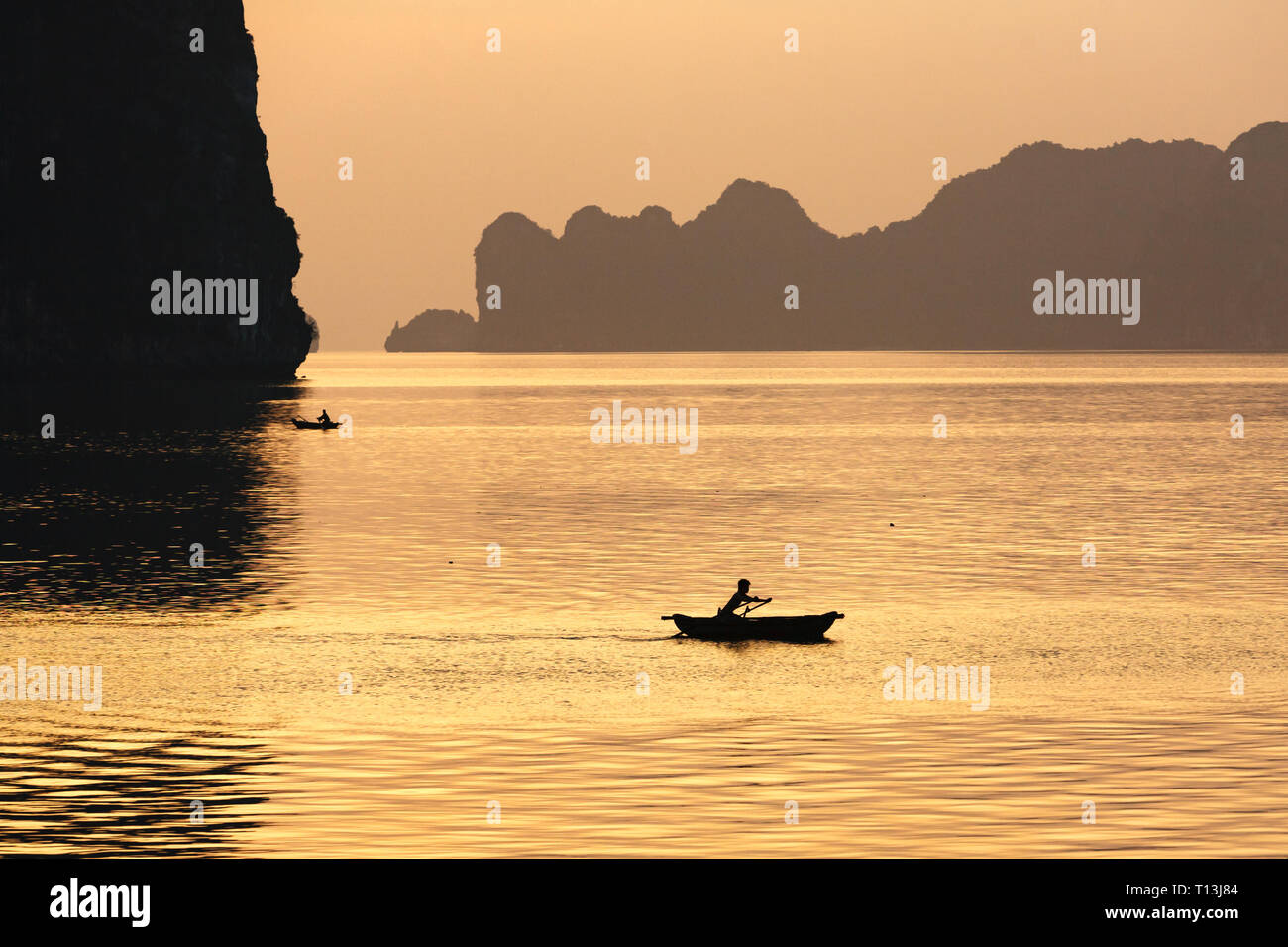 La baie d'Halong dans la lumière dorée du coucher de soleil avec des autochtones dans l'eau des canots crossing Banque D'Images
