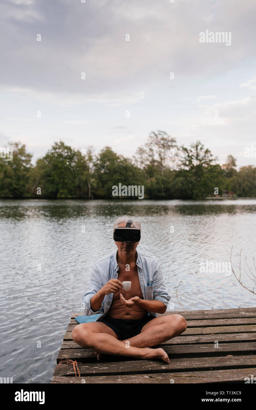 Senior man sitting on jetty at un lac portant des lunettes VR et de boire du café Banque D'Images