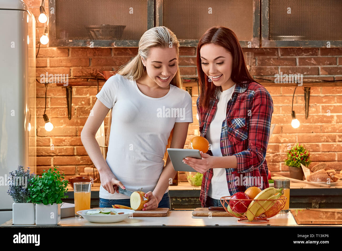 Portrait de deux femmes attrayants, s'amuser, alors que la préparation de salades de fruits. Ils sont pleinement impliqués dans le processus. Fille blonde en T-shirt blanc et son ami brun en chemise à carreaux à la recherche de recettes santé, à l'aide de tablet pc. Ils sont impressionnés par la variété de choix. Vue avant Banque D'Images