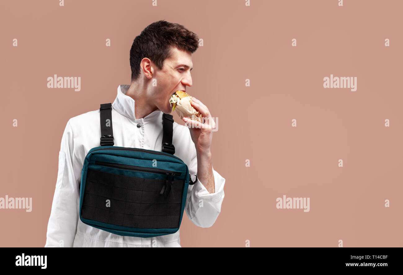 Jeune travailleur homme élégant avec poitrine rig bag est en train de manger un délicieux burger juteux sur fond blanc isolé sur le fond de couleur Banque D'Images