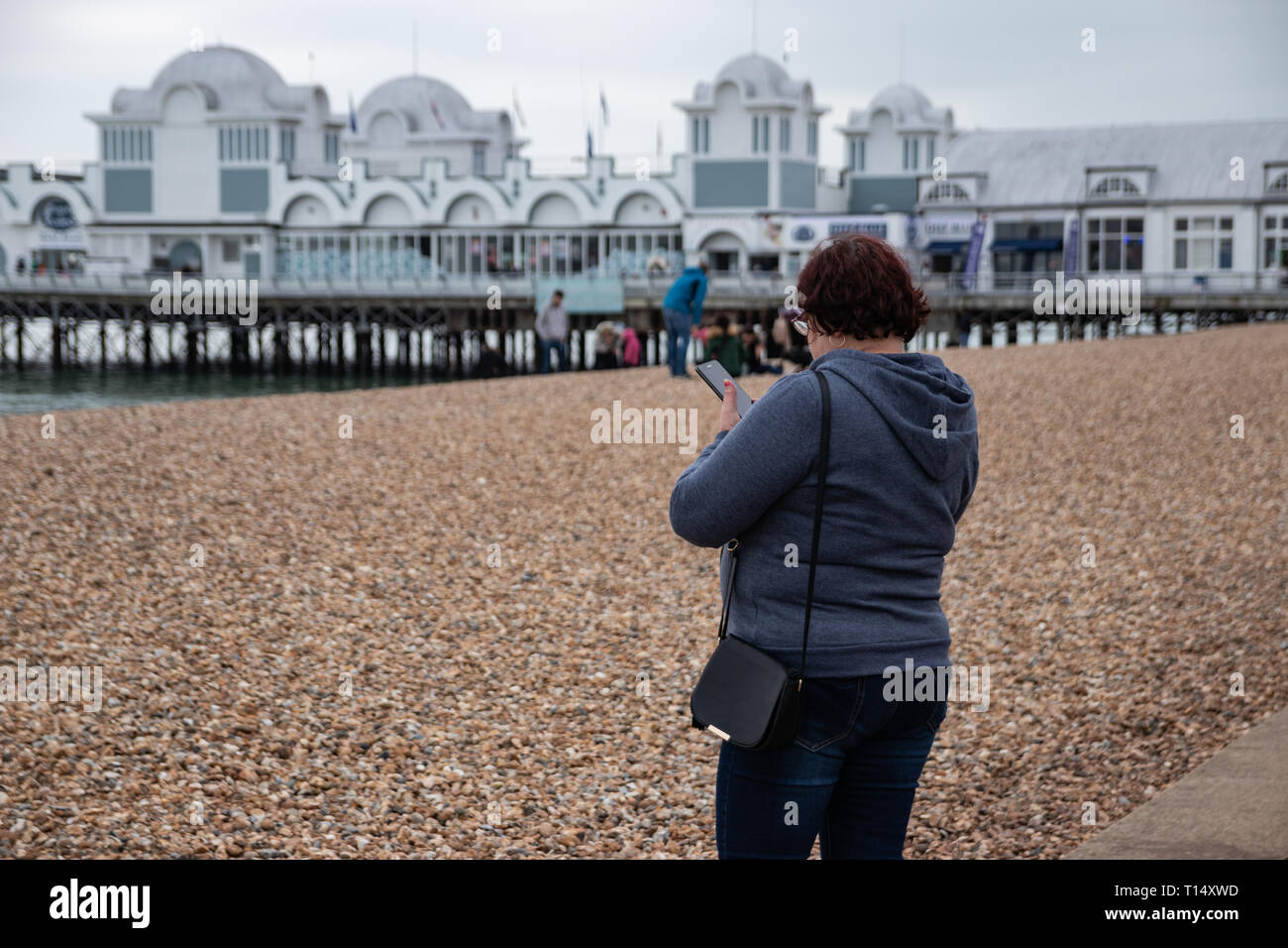 Femme d'âge moyen à l'aide de téléphone mobile à la plage avec jetée dans l'arrière-plan Banque D'Images