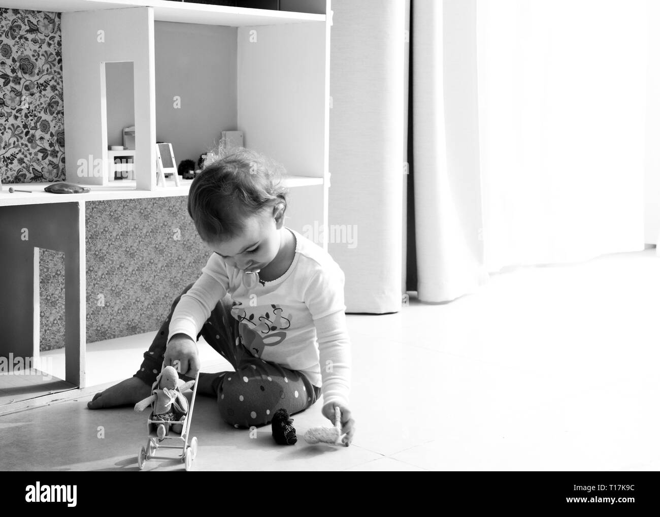 image rétro-éclairée d'un enfant qui joue avec une maison de poupée et des jouets animaux en peluche, assis sur un sol carrelé, seul à la maison avec une sucette dans la bouche. Banque D'Images