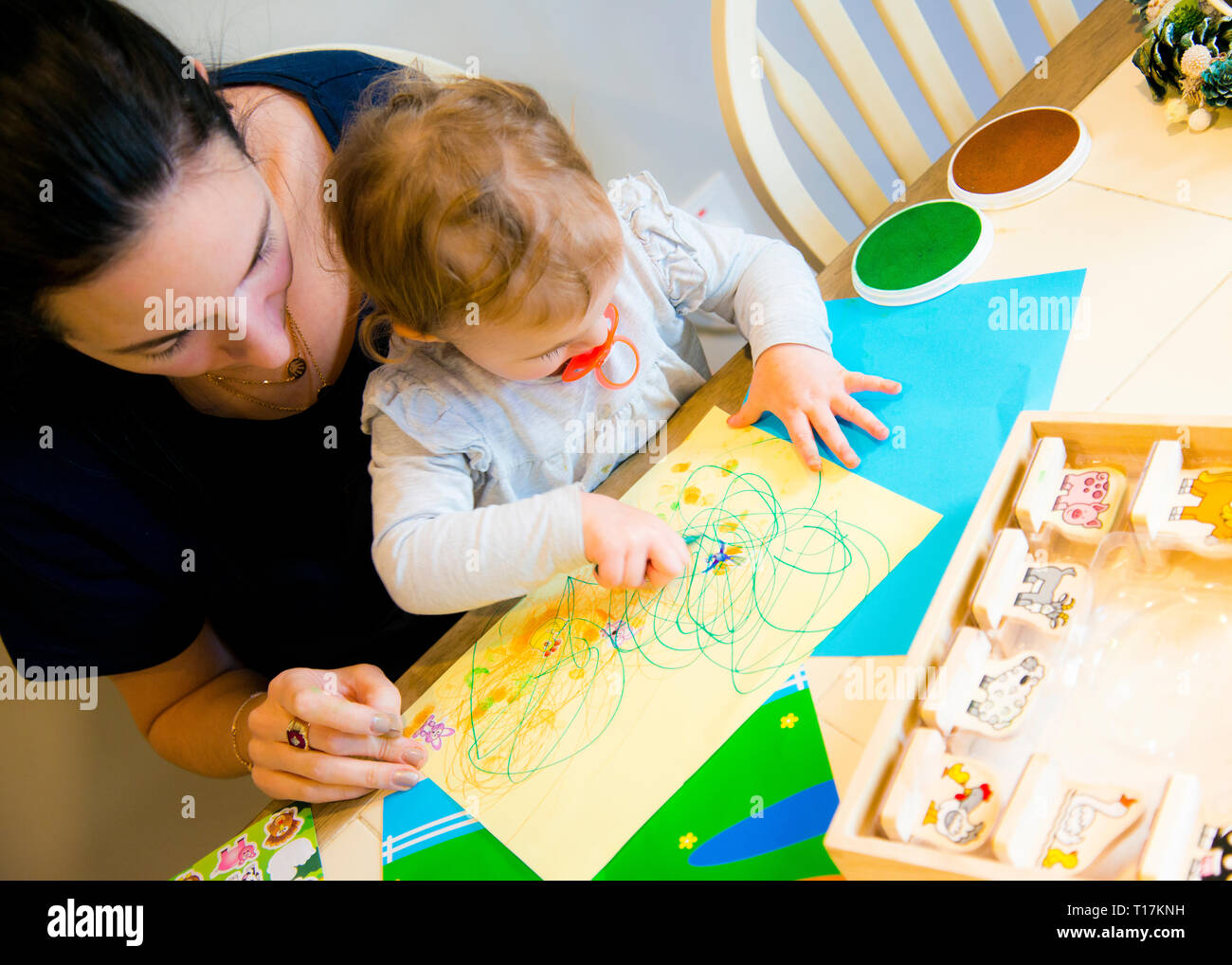 Vue en grand angle de la mère aidant la jeune fille à exprimer son art sur papier trop coloré, assis à la table de cuisine. Banque D'Images