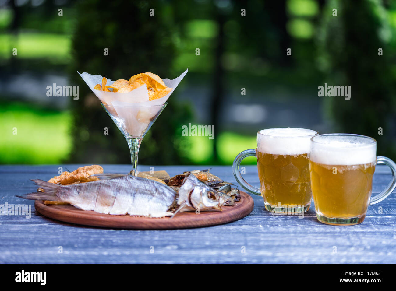 Des en-cas pour la bière et deux chopes de bière. Poisson salé, séché, encornet anneaux et chips de pomme de terre, sur la planche à découper et vert fond d'été Banque D'Images