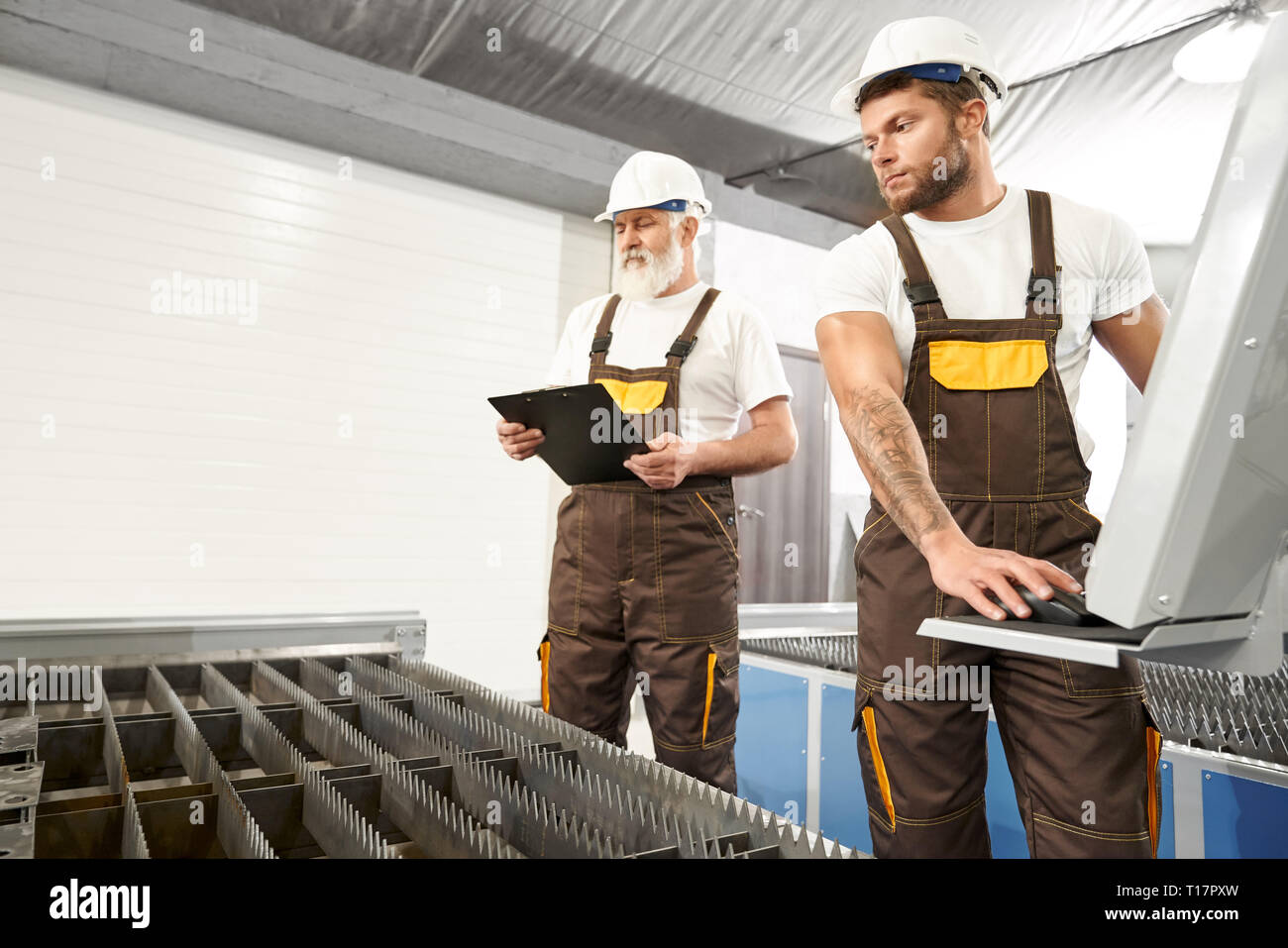 Les ingénieurs expérimentés de l'usine portant des casques blancs et de t shirts, brown une combinaison. Vieil homme noir, dossier holding laser observation plasma cutter, jeune homme machine informatisé d'exploitation. Banque D'Images