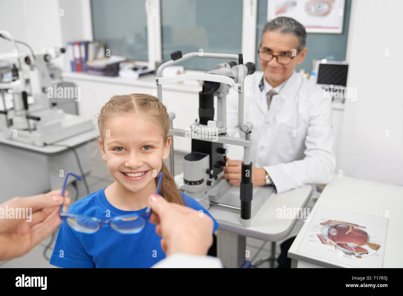 Pretty girl smiling and looking at camera. Les mains des femmes pour donner le choix de lunettes bleu les patients. Médecin Optométriste assis à table avec lampe à fente dans la salle médicale de la clinique. Banque D'Images