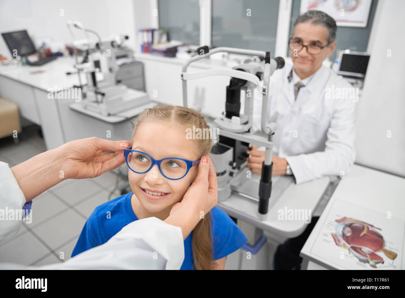 Mains de médecin portant des lunettes bleu sur patient. Cute girl smiling. Optométriste professionnel travaillant à table avec équipement médical spécial pour l'examen de la vue dans la salle médicale de la clinique. Banque D'Images