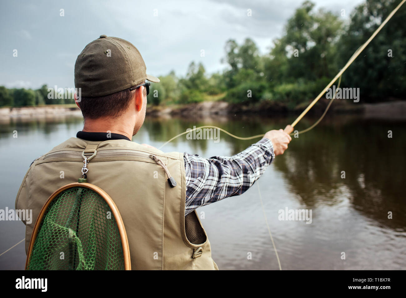 Guy est debout et à tout droit sur l'eau et les arbres. Il porte gilet, casquette et chemise. Guy a également filet de pêche derrière son dos. L'homme détient mouche Banque D'Images