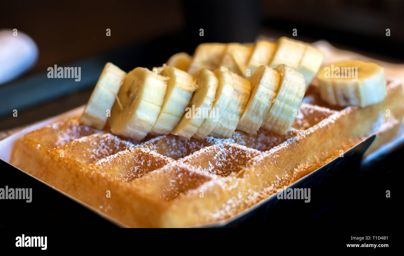 Gaufre Belge carré frais coupés avec garniture de bananes et de sucre en poudre pour le petit déjeuner sur fond sombre. Banque D'Images