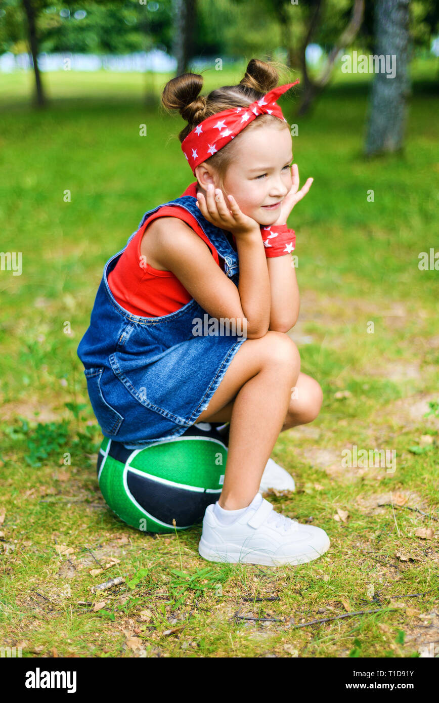 Belle petite girl smiling in denim salopettes red t-shirt, hairstyle. l'enfant est assis sur la balle Banque D'Images