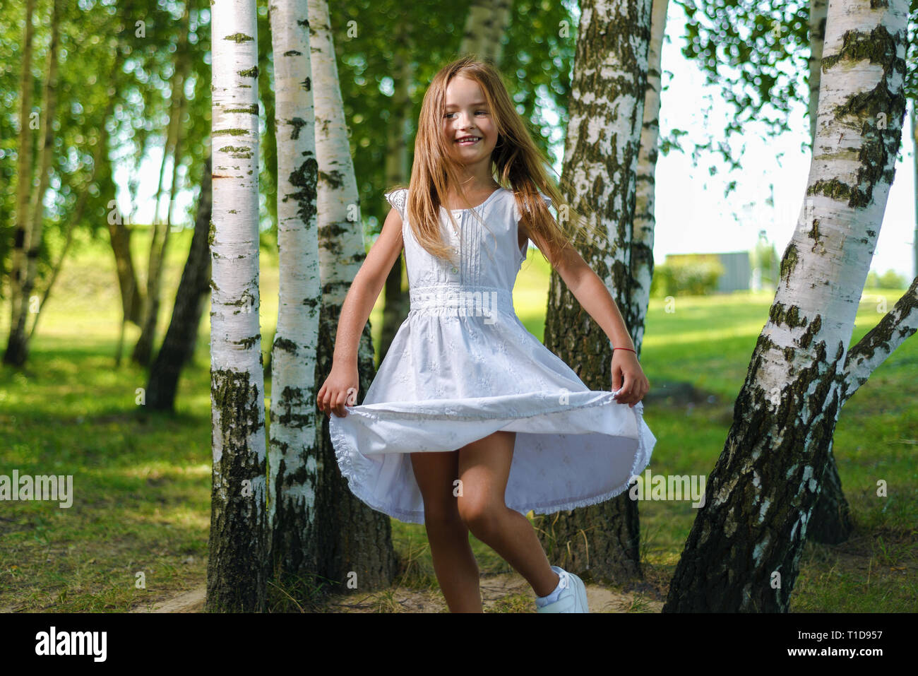 Jolie petite fille en robe blanche et s'enroule et danse entre les arbres du parc Banque D'Images