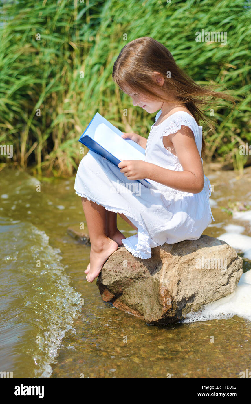 Petite fille en robe blanche est assis sur un rocher près de la rivière aux beaux jours. Heureux enfant lit livre et sourit Banque D'Images