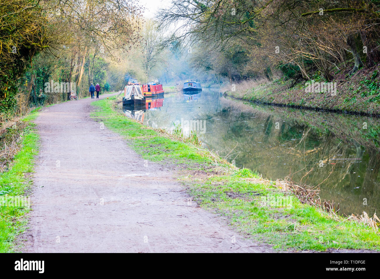 Un couple en train de marcher le long du chemin de halage du canal de Kennet et Avon passant houseboats avec une brume des cheminées des bateaux par un beau jour de printemps. Banque D'Images