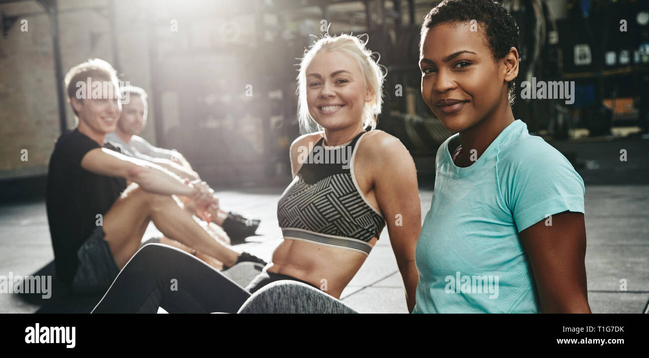 Deux jeunes femmes de sportswear smiling lors d'un entretien avec un couple d'amis sur le plancher d'une salle de sport après une séance Banque D'Images