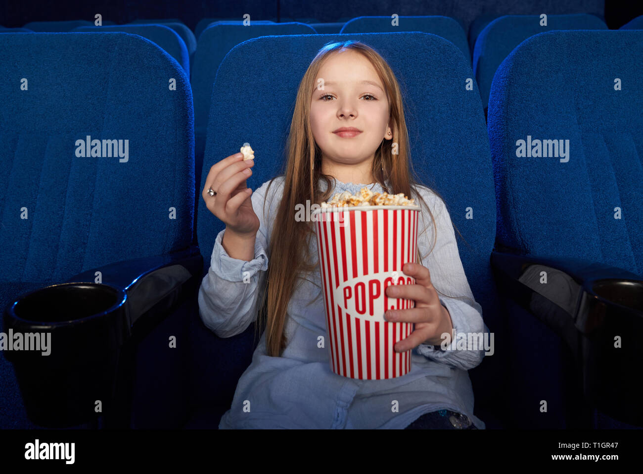 Beautiful girl looking at camera, holding grand seau de popcorn savoureux. Joli gosse assis en bleu foncé chaise confortable en cinéma, bénéficiant d'animation. Banque D'Images