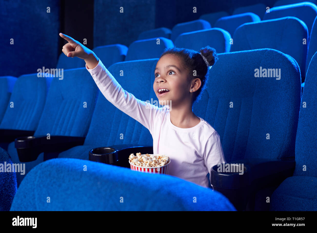 Fasciné girl looking at écran avec choqué face, pointant avec le doigt. Belle enfant africain regarder film en cinéma, holding seau de maîs éclaté. Banque D'Images