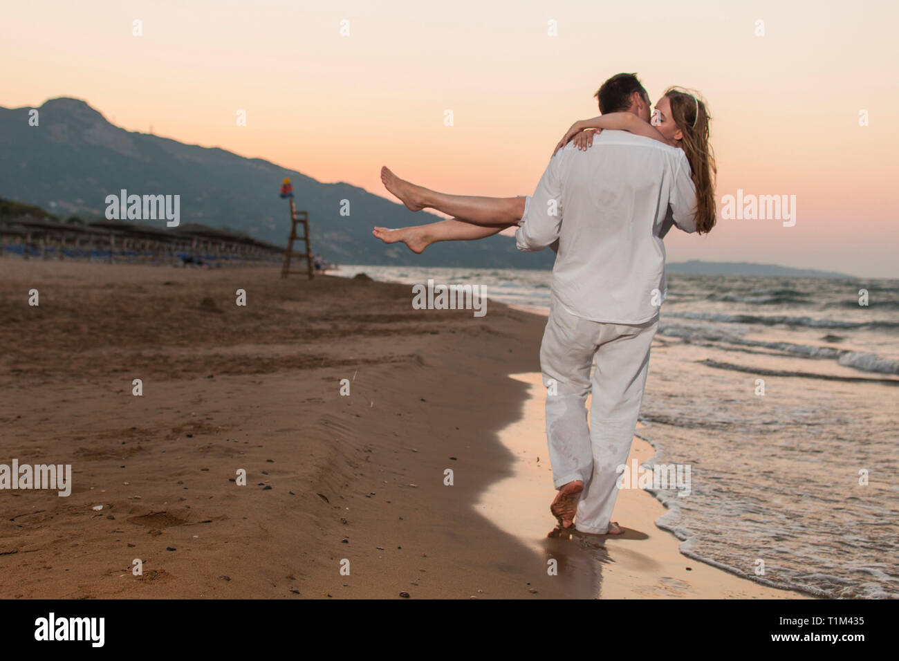 Vue romantique de l'homme qui porte sa bien-aimée femme en marchant à travers les mains de sable et ils s'embrassent. Magnifique paysage de bord de mer au coucher du soleil. Banque D'Images