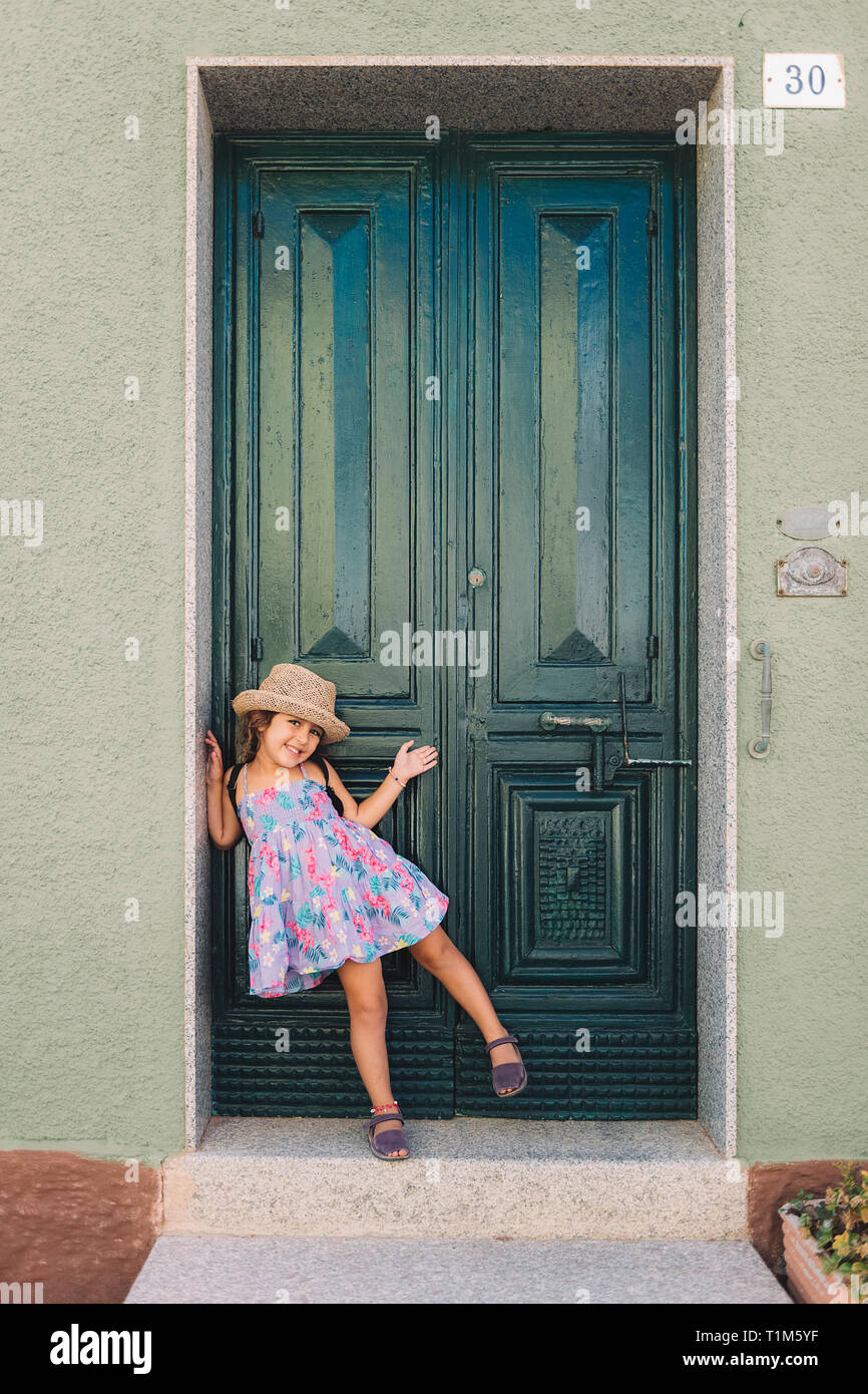 Petite fille pose amusé et souriant en face de la porte de sa maison, elle  porte une robe d'été lilas de fleurs et un chapeau de paille, vertical imag  Photo Stock -