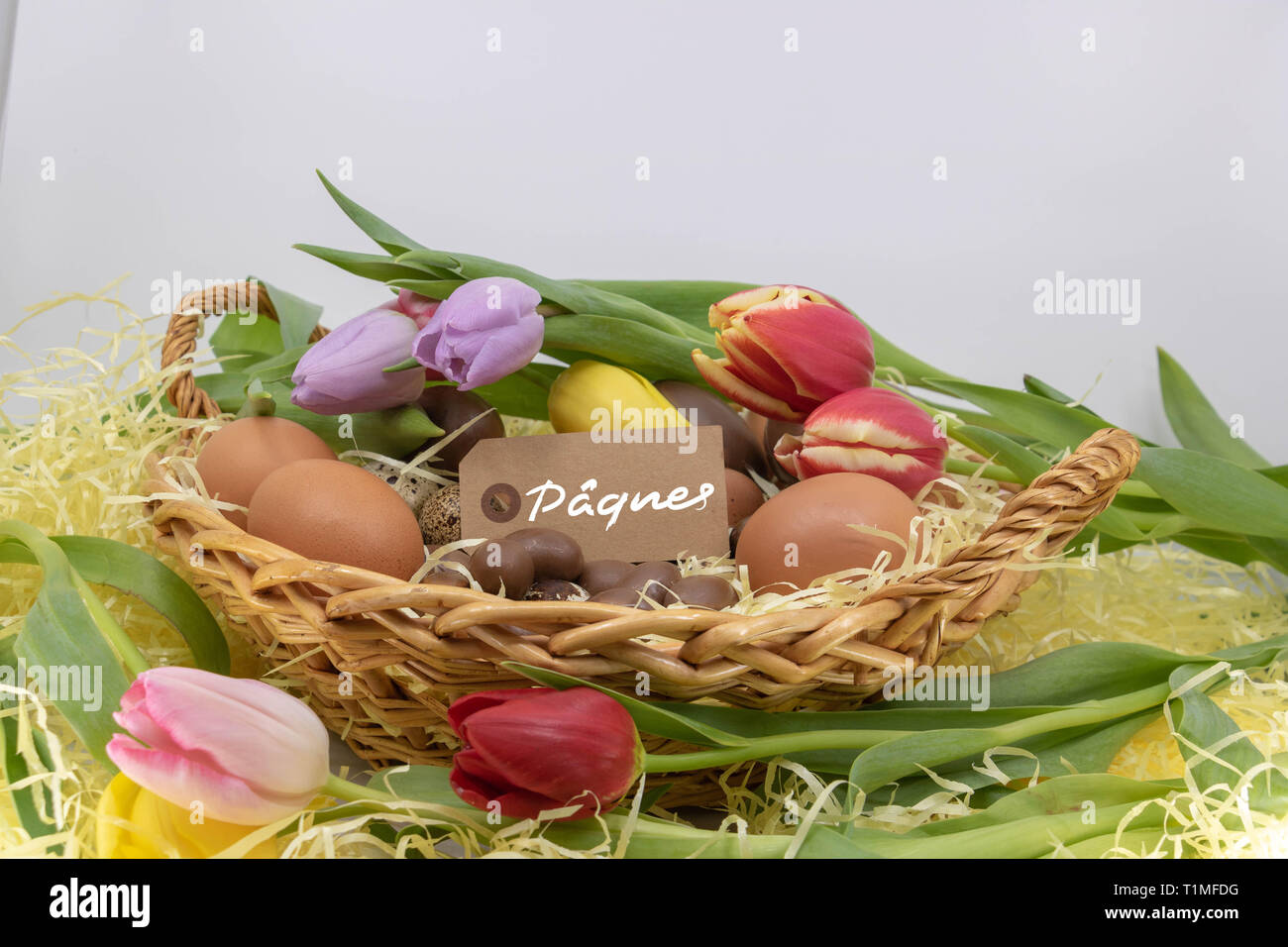 Pâques Pâques est écrit en français sur une étiquette dans un panier rempli d'oeufs de poule, caille, chocolat ainsi que les tulipes Banque D'Images