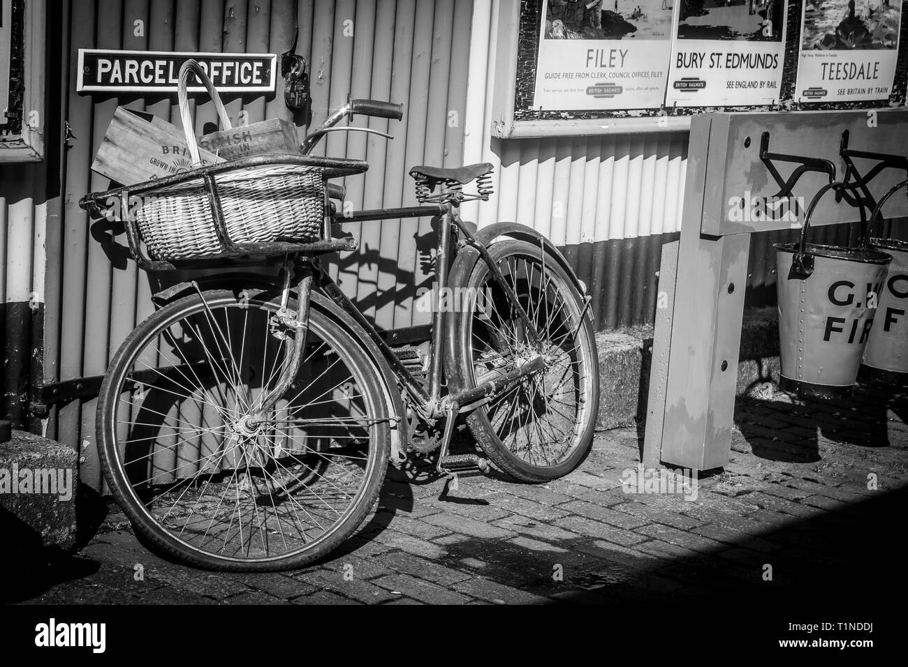Gros plan monochrome de la vieille bicyclette, vélo, panier britannique vintage sur le devant de la réception, contre le bureau de colis sur la plate-forme de la gare du patrimoine britannique. Banque D'Images