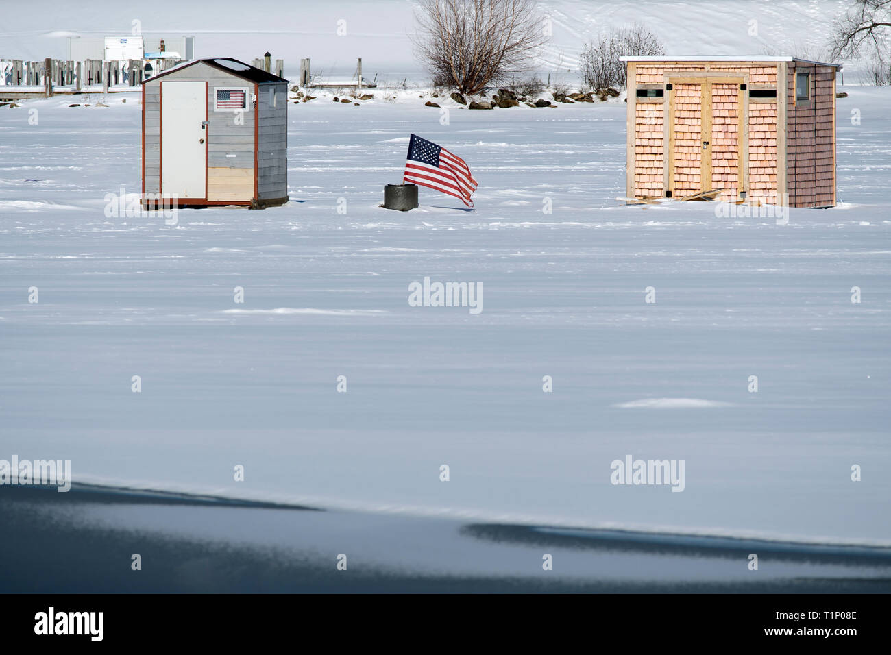 Drapeau américain affiche entre deux cabanes de pêche sur glace près de la rive du lac que la glace commence à fondre sur le rivage dans le New Hampshire. Banque D'Images