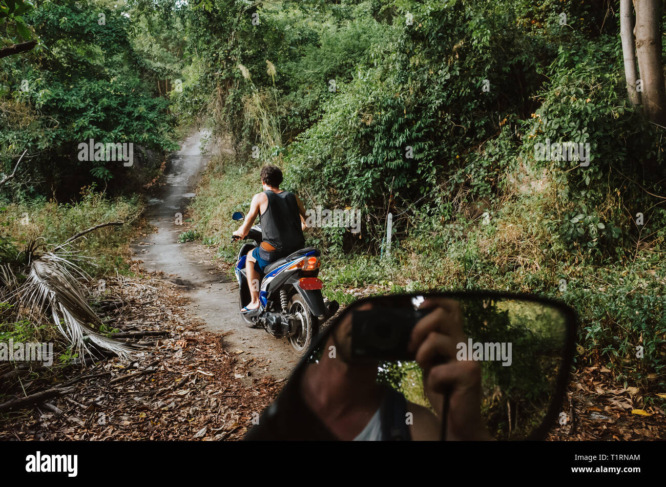 Jeune homme sur un scooter roulant sur un chemin dans la jungle alors qu'un autre homme peut être vu à travers le miroir arrière de prendre une photo de lui Banque D'Images