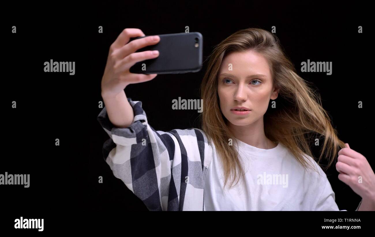 Portrait de jeune fille aux longs cheveux caucasiens chemise en faisant des photos à l'aide de selfies smartphone sur fond noir. Banque D'Images
