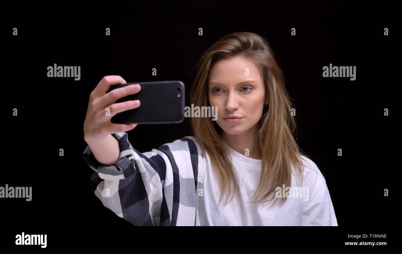 Portrait de la belle jeune fille aux longs cheveux caucasiens chemise en faisant des photos à l'aide de selfies smartphone sur fond noir. Banque D'Images
