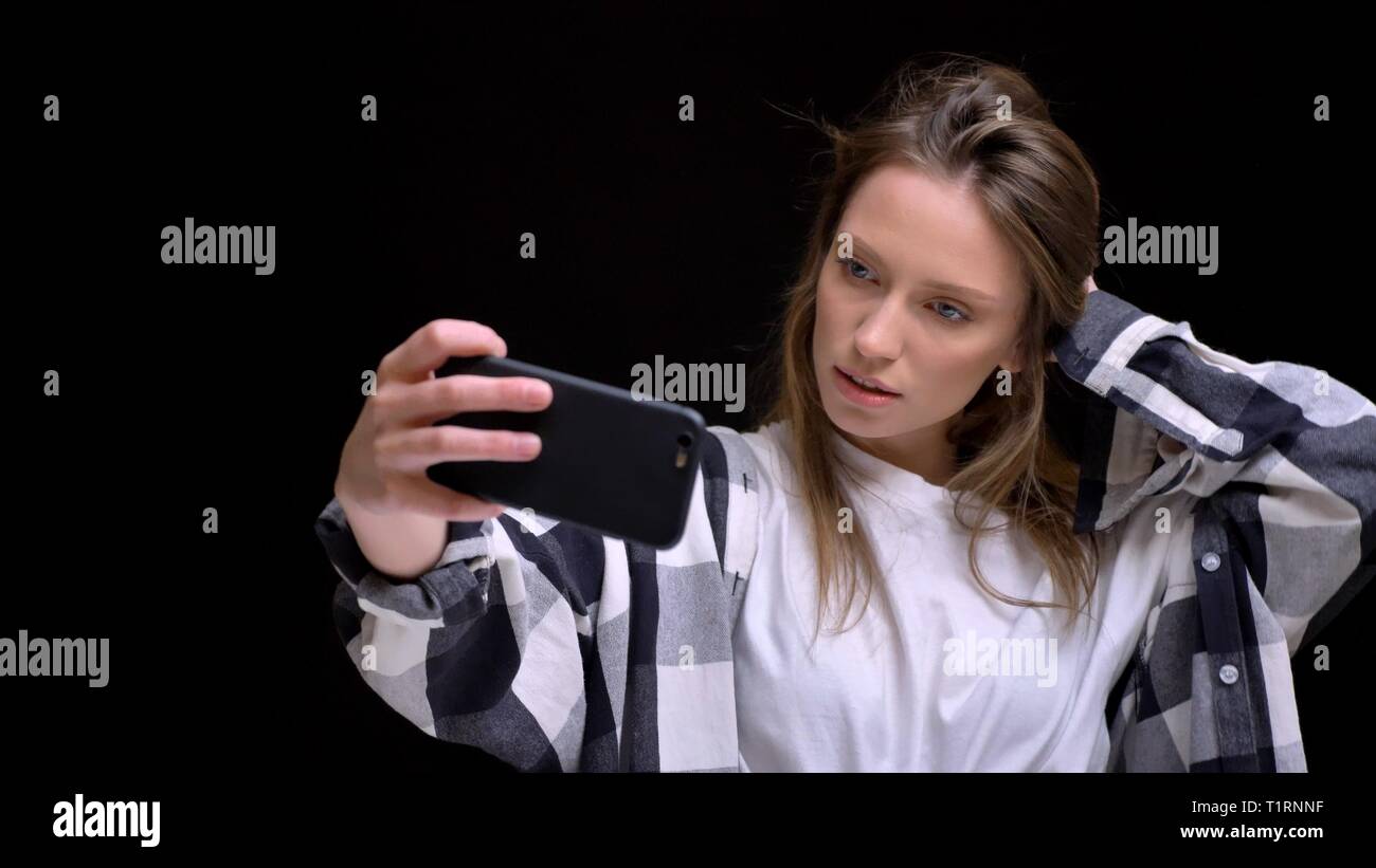 Portrait de jeune fille aux longs cheveux blancs en chemise à carreaux de toucher ses cheveux et faire des photos à l'aide de selfies smartphone sur fond noir. Banque D'Images