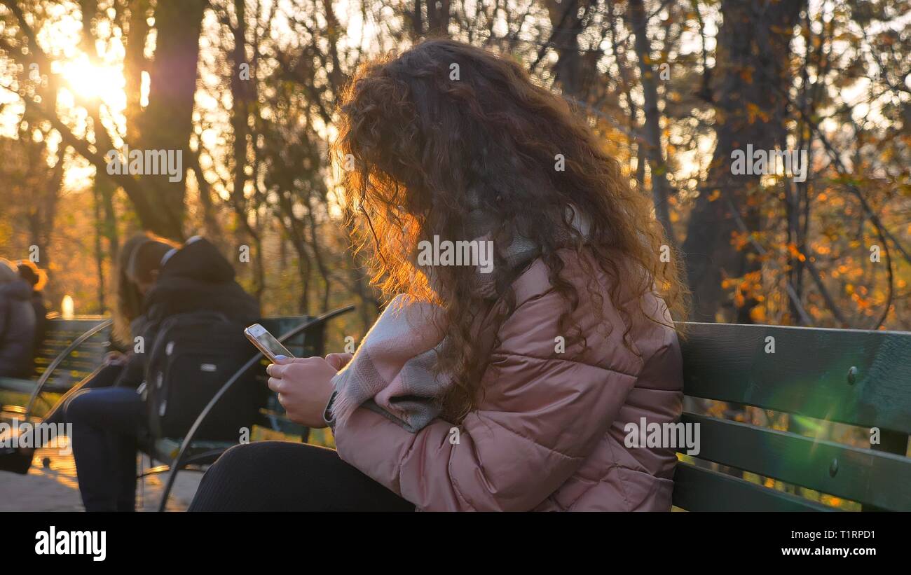 Portrait de jeune fille de race blanche aux cheveux bouclés assis sur un banc et regarder sur smartphone dans le parc d'automne. Banque D'Images