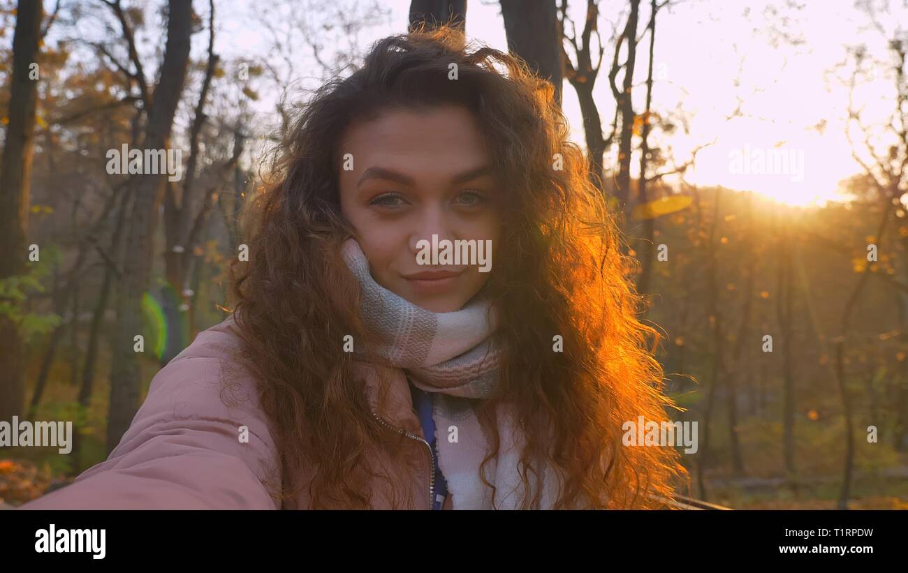 Les selfies photo de jeune fille de race blanche aux cheveux bouclés en souriant et en regardant la caméra avec un clin d il à l'automne dans le parc ensoleillé. Banque D'Images