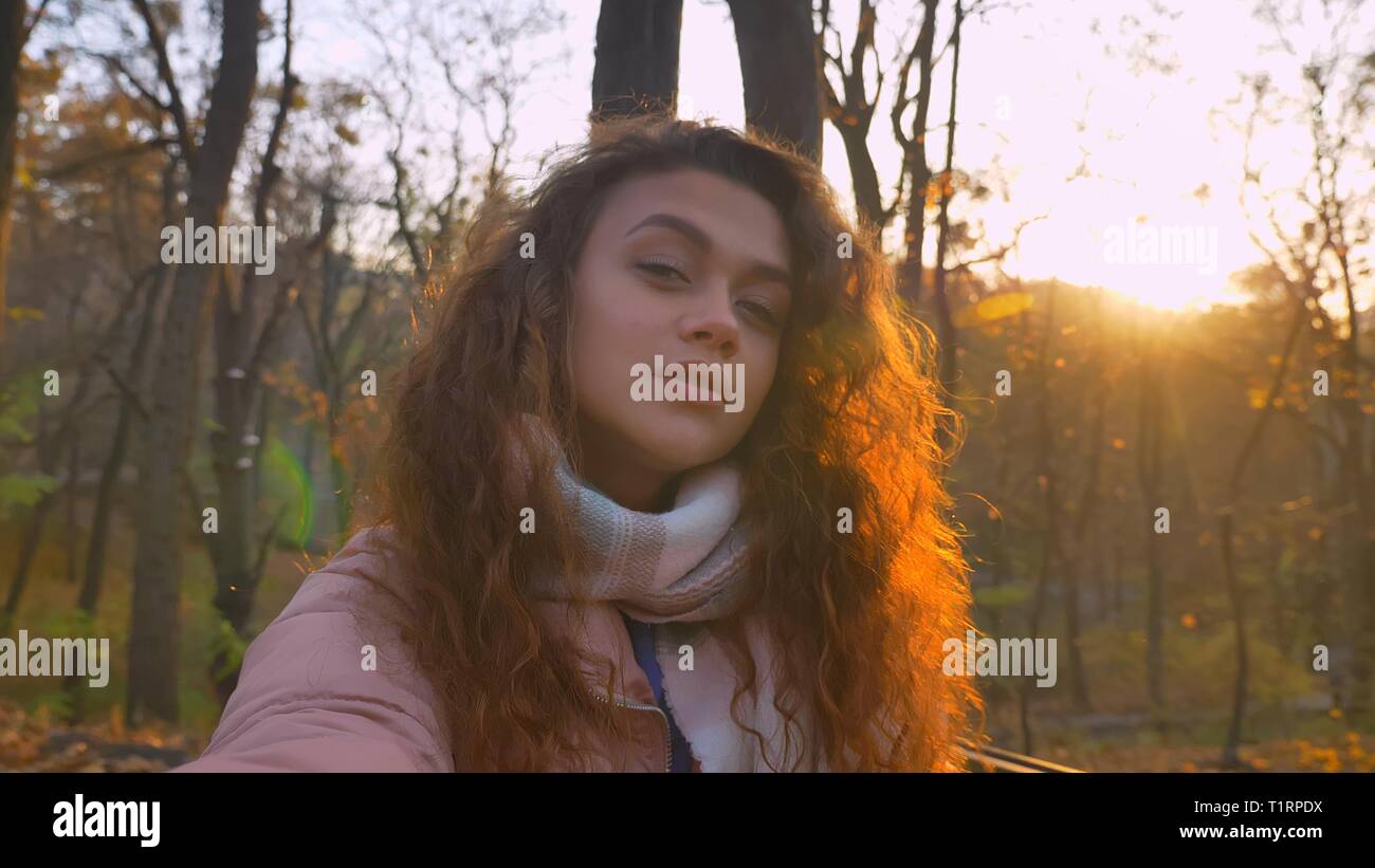 Les selfies photo de curly-haired woman regarder sérieusement en caméra et de flirter avec elle dans le parc d'automne ensoleillé. Banque D'Images