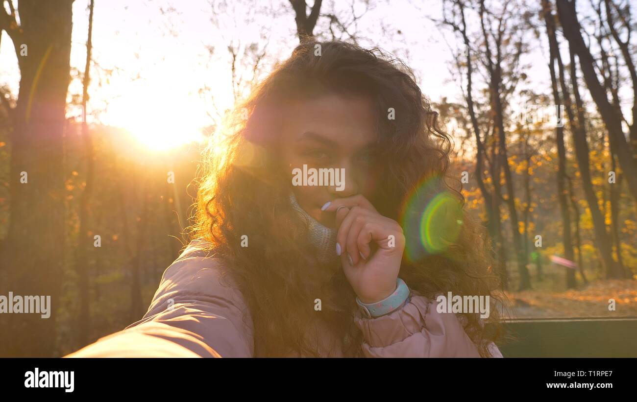 Les selfies photo de jolie fille de race blanche aux cheveux bouclés avec les doigts sous le nez de l'appareil photo en regardant sous le soleil de parc d'automne. Banque D'Images