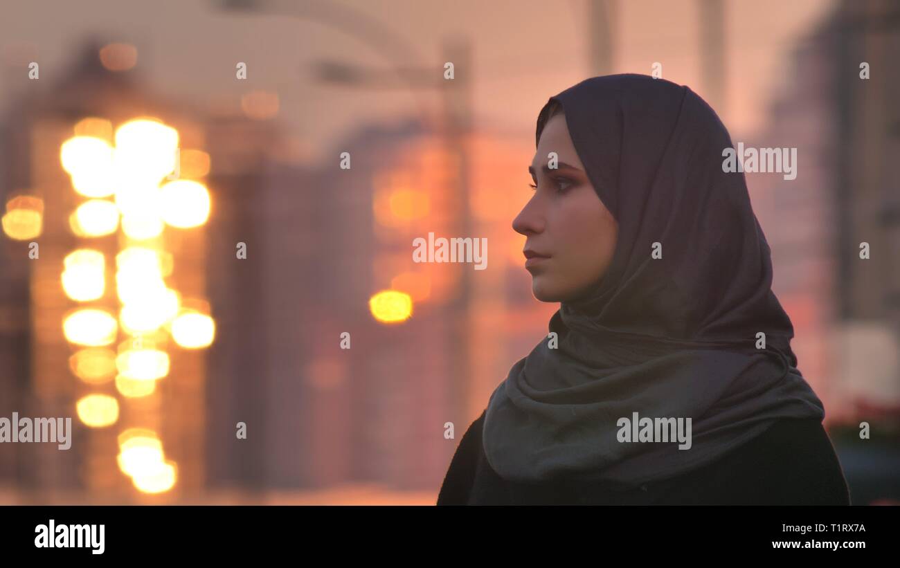 Closeup portrait of young pretty femme en hijab et tournant à la recherche sur le côté avec les bâtiments lumineux sur l'arrière-plan Banque D'Images