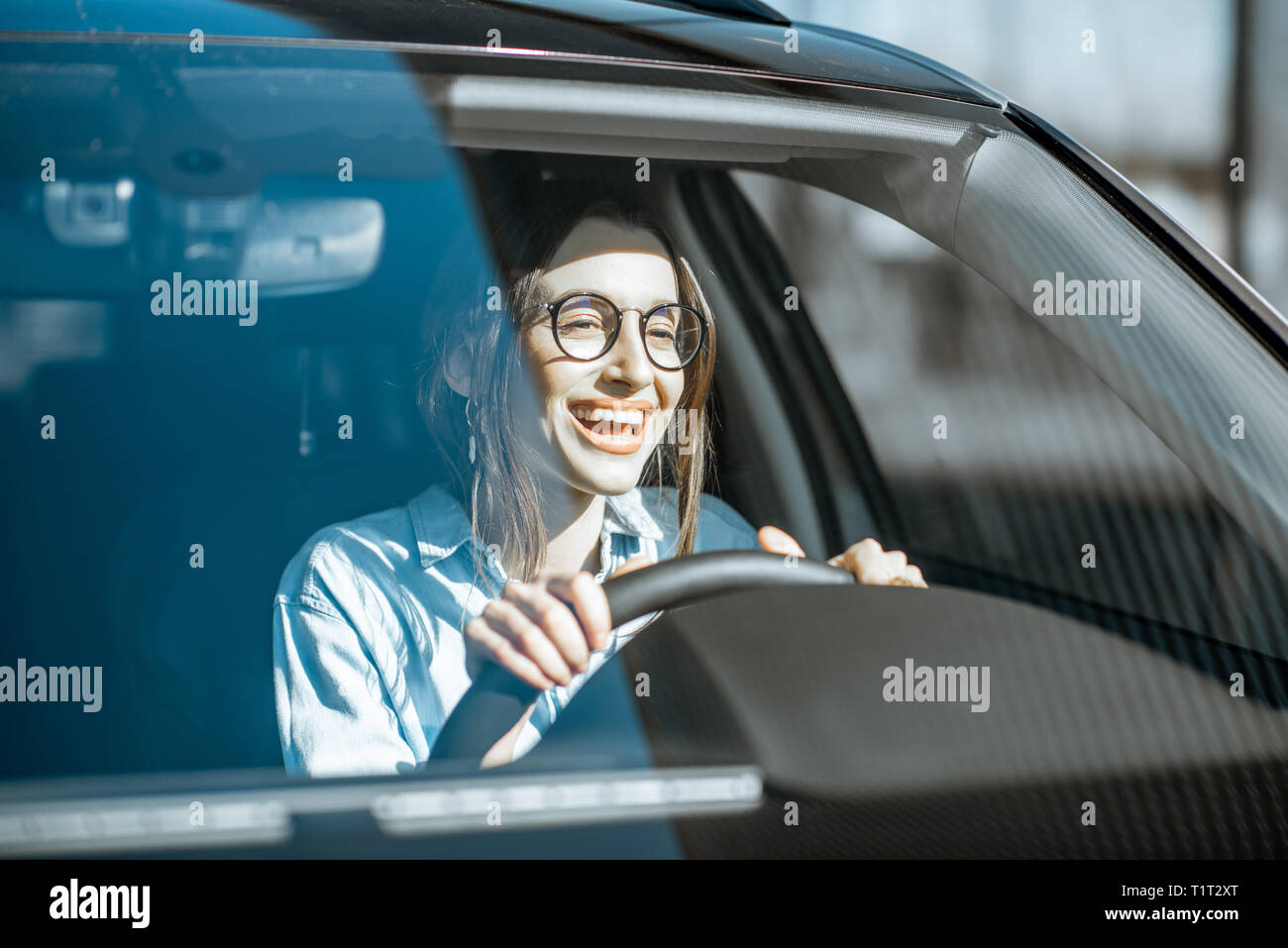 Jeune femme heureuse et conduire une voiture de luxe, vue de face avec la lumière du soleil à travers le pare-brise Banque D'Images