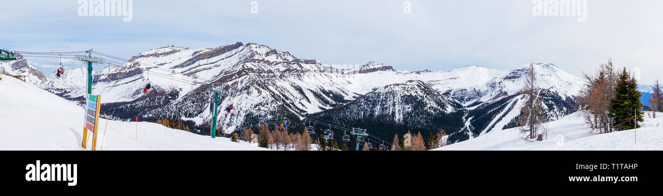 Panorama grand angle de skieurs et planchistes sur télésiège remontant une pente de ski dans la montagne enneigée (éventail des Rocheuses canadiennes au lac Louise. Banque D'Images