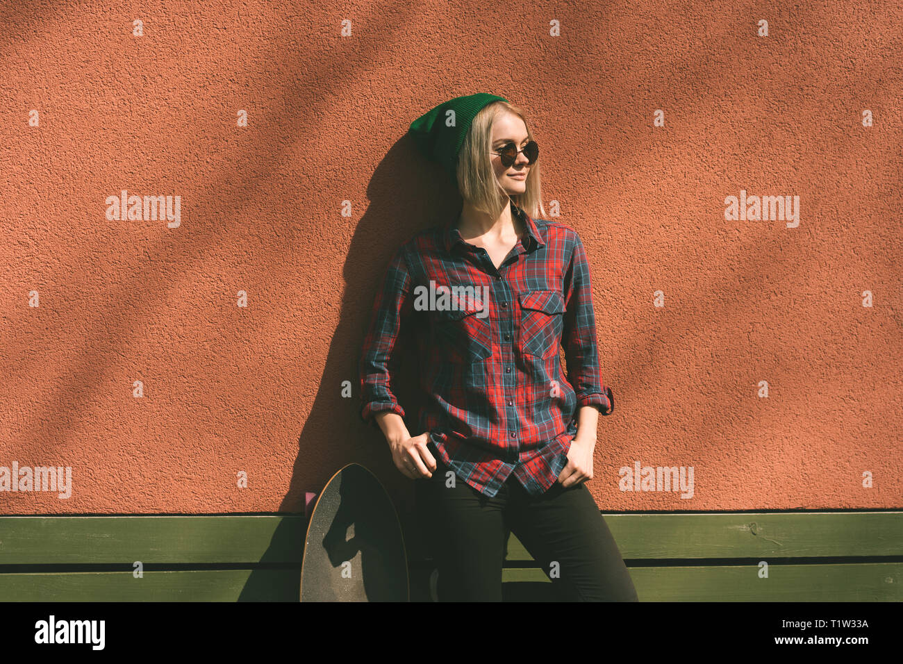 Hipster girl blonde en chemise à carreaux et un chapeau vert avec une planche à roulettes sur un fond de corail. Banque D'Images