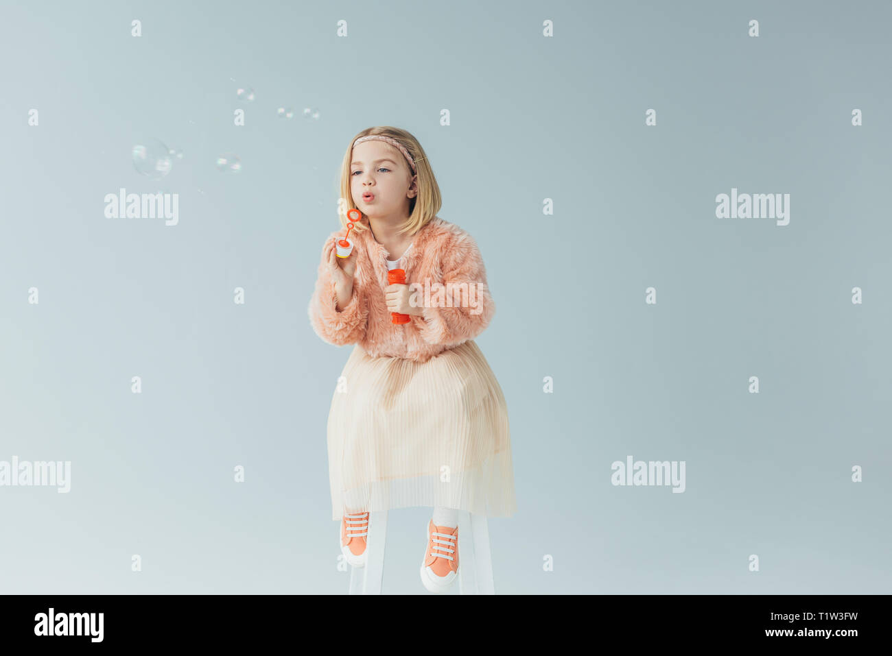 Adorable gamine dans un manteau en fausse fourrure et jupe assise sur une chaise haute et soufflant des bulles de savon isolé sur gray Banque D'Images