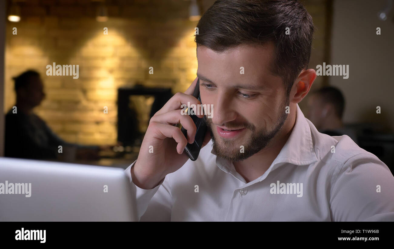 Libre de tirer des profils caucasian businessman typing sur l'ordinateur portable et d'avoir une conversation informelle sur le téléphone en souriant gaiement à l'intérieur dans le bureau Banque D'Images