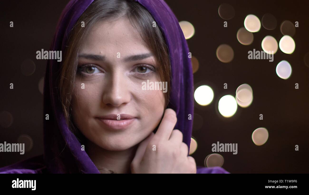 Close-up portrait of beautiful young brunette girl in hood regarder avec calme sourire dans le fond de l'appareil photo Lumières floues. Banque D'Images