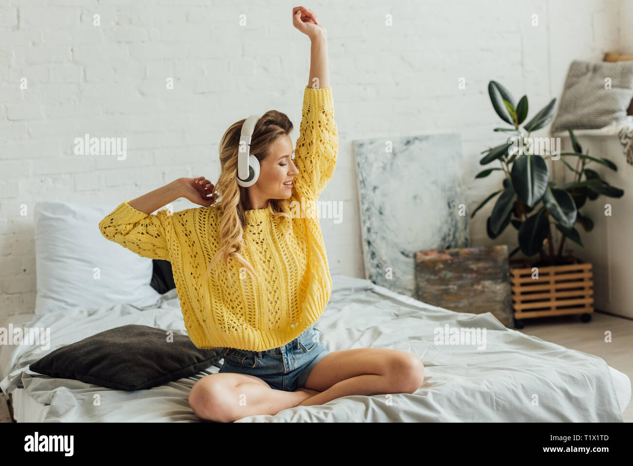 Beautiful smiling young woman in headphones assis sur le lit, s'étirant et en écoutant la musique à la maison Banque D'Images