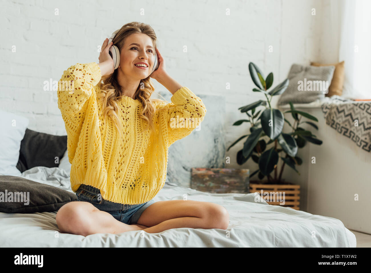 Beautiful happy young woman in headphones assis sur le lit et écoute la musique à la maison Banque D'Images